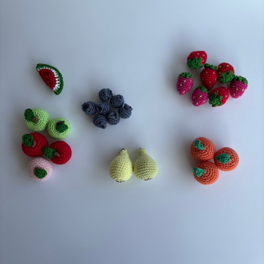 Verschiedene gehäkelte Obstsorten aus Baumwollgarn als Spielzeug für die Kinderküche, darunter Äpfel, Birnen, Orangen, Erdbeeren, Blaubeeren und ein Stück Melone – handgefertigtes Amigurumi-Spielset für Kinder ab 0 Jahren.