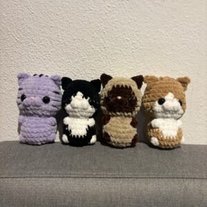 Vier gehäkelte Katzen-Amigurumi in verschiedenen Farben (lila, schwarz-weiß, beige-braun, hellbraun-weiß) stehen nebeneinander auf einem grauen Sofa.