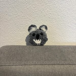 ehäkelter Koala-Amigurumi aus grauem Plüschgarn mit schwarzen Sicherheitsaugen, großer schwarzer Nase, weißem Bauchdetail und flauschigen Ohrpuscheln, sitzend auf grauem Sofa.