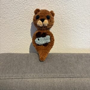 Gehäkelter Otter-Amigurumi aus braunem Plüschgarn mit schwarzen Sicherheitsaugen, hellem Schnäuzchen und kleinem Fisch am Bauch, sitzend auf grauem Sofa.