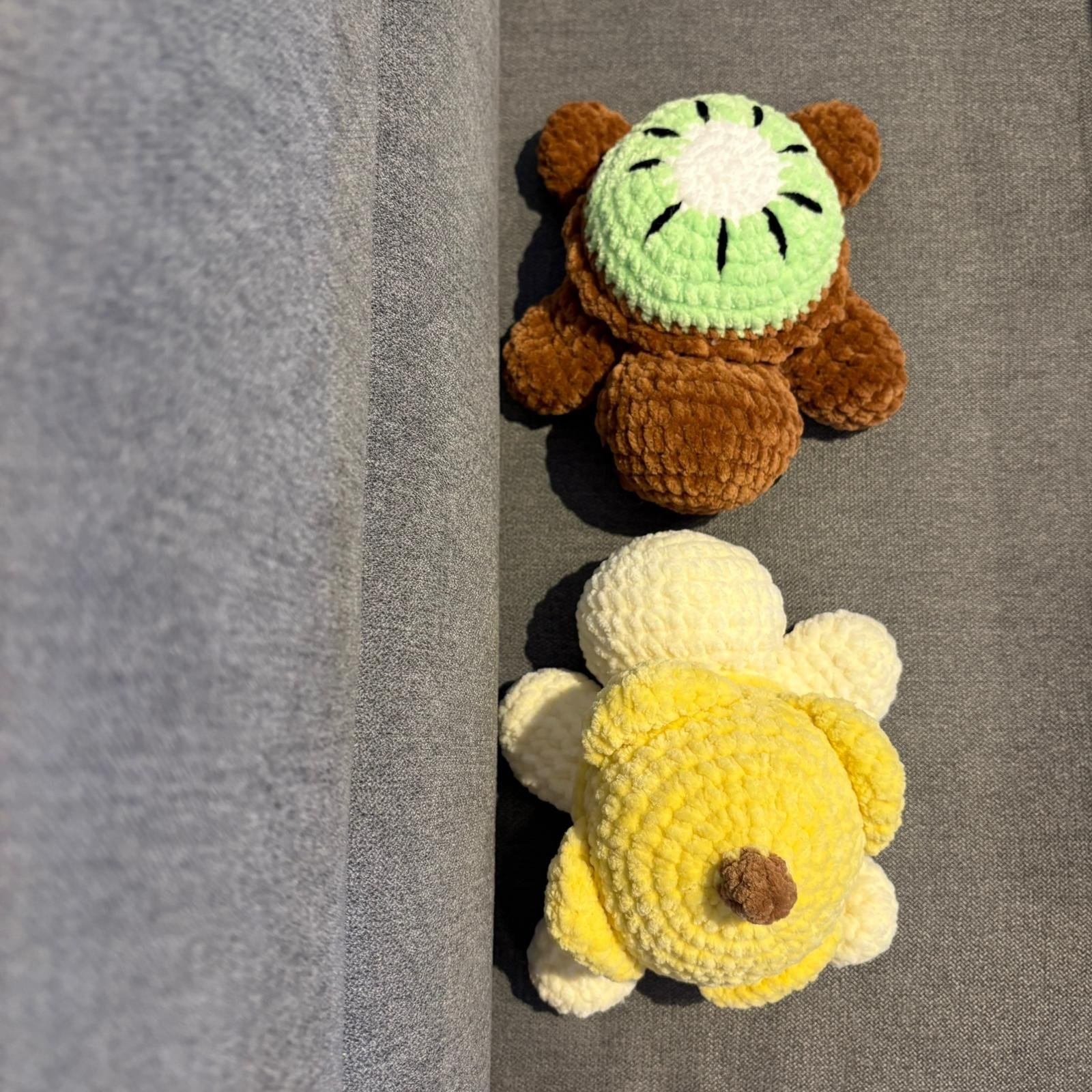 Draufsicht auf zwei gehäkelte Schildkröten: braun mit Kiwi-Panzer und weiß-gelb mit Bananen-Panzer auf grauem Sofa.