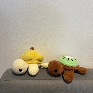 Zwei gehäkelte Schildkröten-Amigurumi liegen nebeneinander auf einem grauen Sofa: links Bananen-Design, rechts Kiwi-Design.