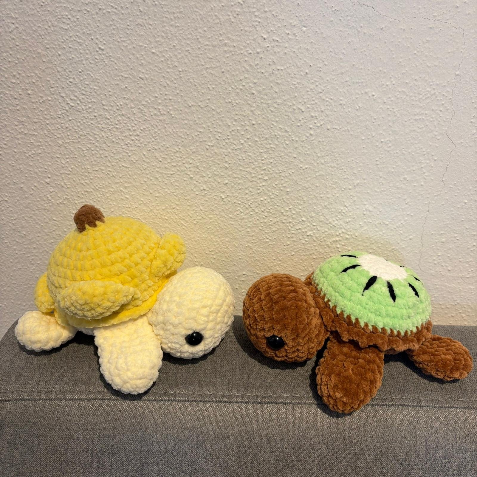 Seitliche Ansicht der gehäkelten Frucht-Schildkröten auf grauem Sofa: gelb-weiß (Bananen) neben braun-grün (Kiwi).