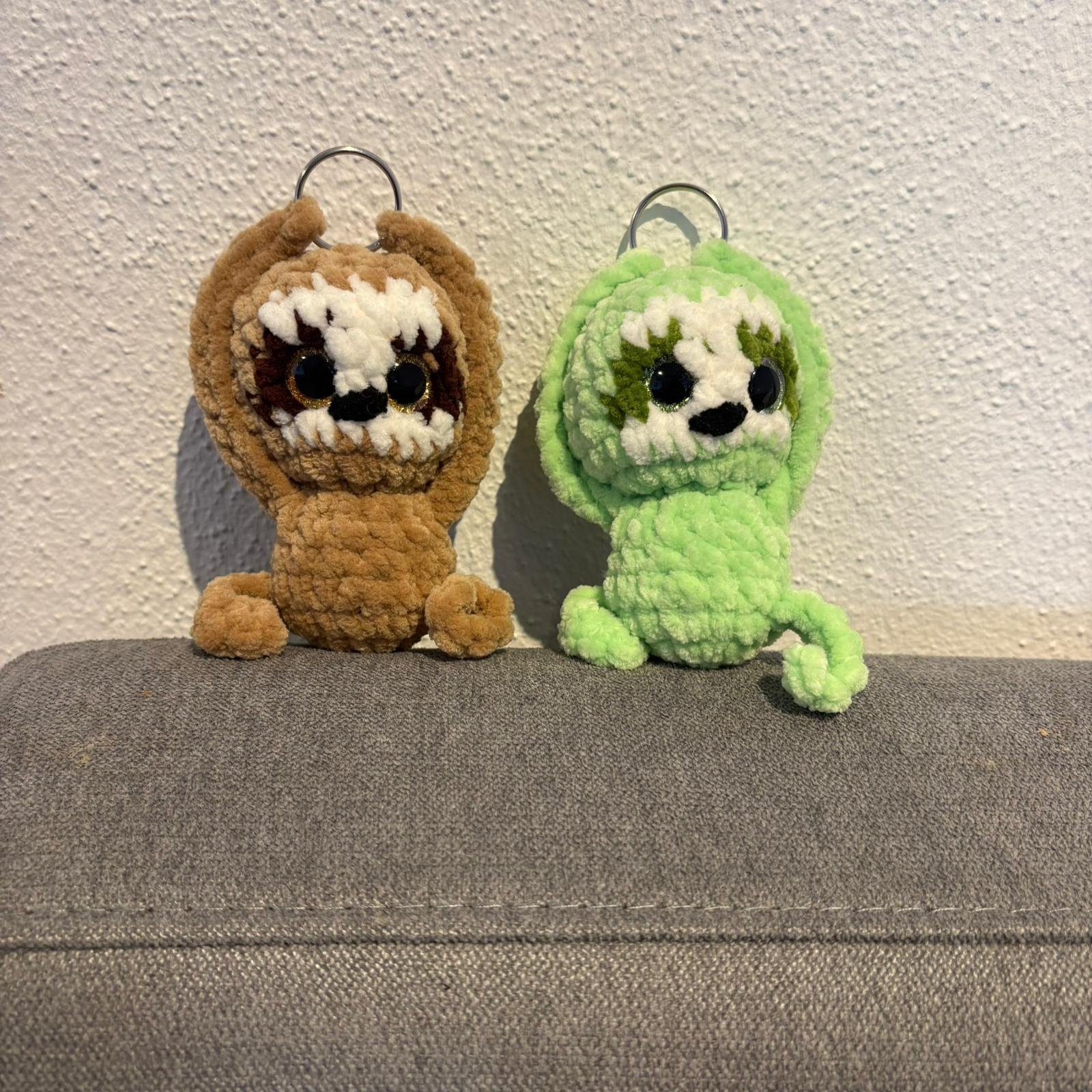 Zwei gehäkelte Faultier-Schlüsselanhänger (braun und grün) stehen aufrecht nebeneinander, mit Schlüsselring oben am Kopf und süßem Faultier-Gesicht.