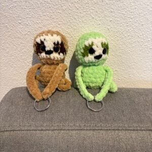 Zwei gehäkelte Faultier-Schlüsselanhänger (braun und grün) mit großen Glitzeraugen sitzen auf einem grauen Sofa, die Arme enden in Schlüsselringen.