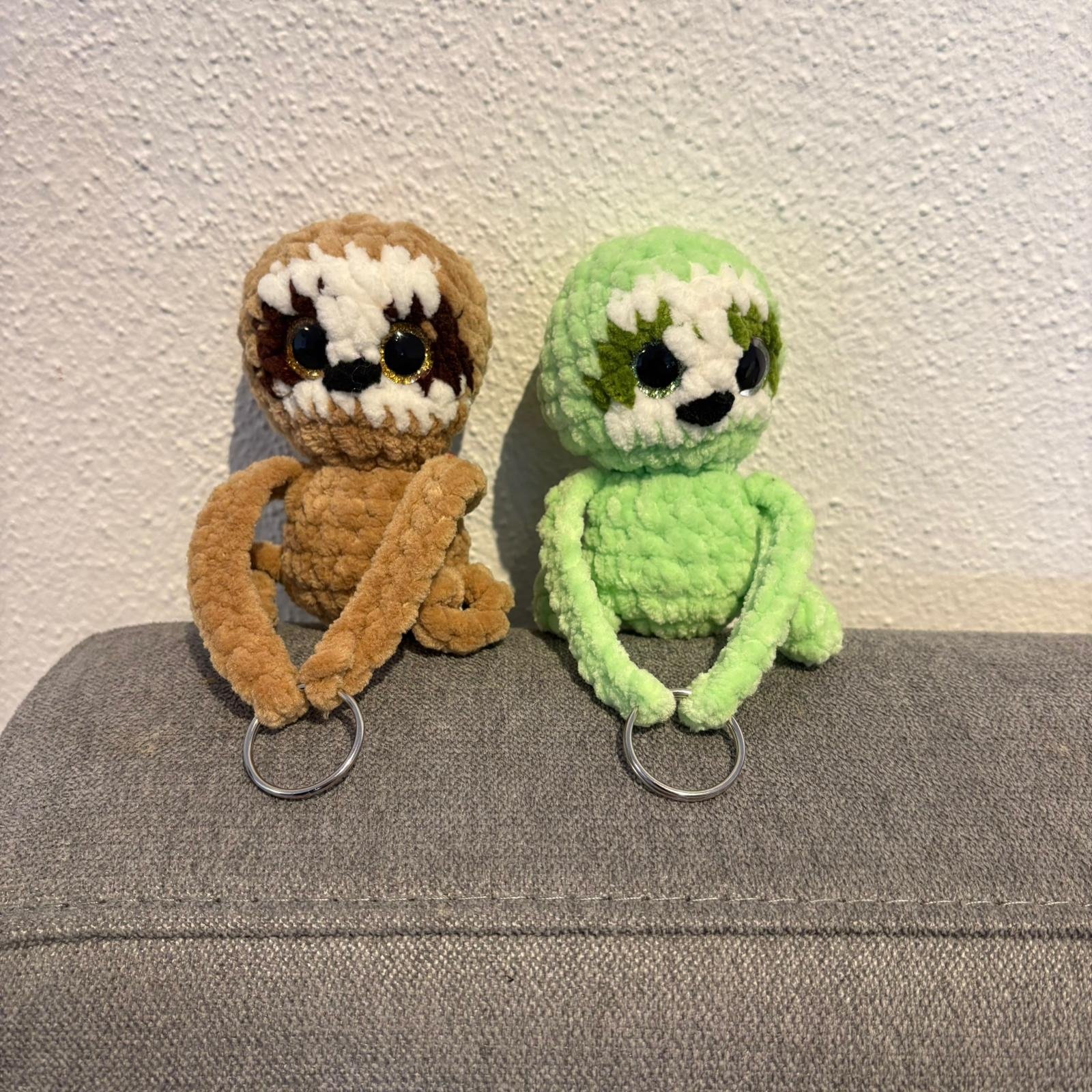 Zwei gehäkelte Faultier-Schlüsselanhänger (braun und grün) mit großen Glitzeraugen sitzen auf einem grauen Sofa, die Arme enden in Schlüsselringen.