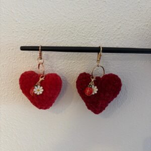 Zwei gehäkelte Herz-Schlüsselanhänger in Rot und Dunkelrot hängen an einer schwarzen Stange, jeweils mit Marienkäfer- und Gänseblümchen-Anhänger.