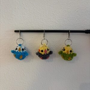 Drei gehäkelte Wellensittich-Schlüsselanhänger (blau-weiß, gelb-grau und gelb-grün) hängen an einer schwarzen Stange, jeweils mit Metall-Schlüsselring.