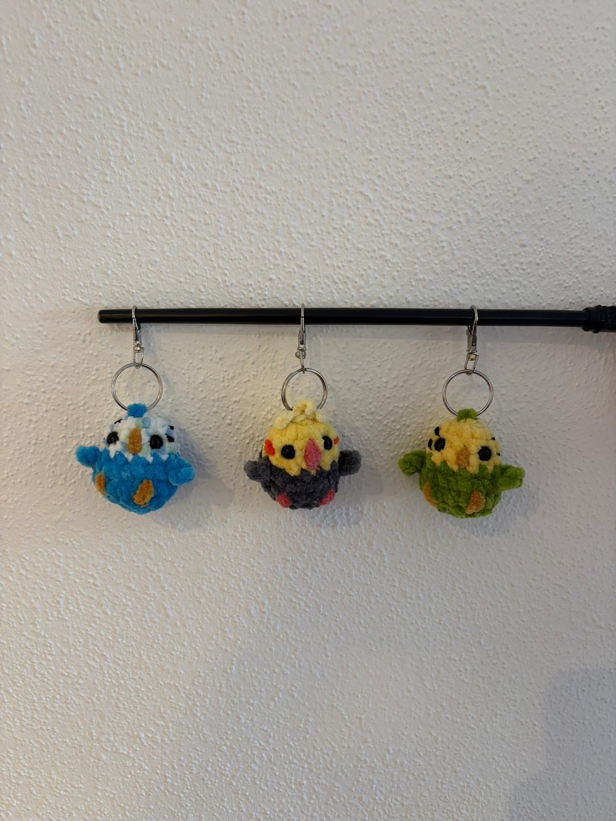 Drei gehäkelte Wellensittich-Schlüsselanhänger (blau-weiß, gelb-grau und gelb-grün) hängen an einer schwarzen Stange, jeweils mit Metall-Schlüsselring.
