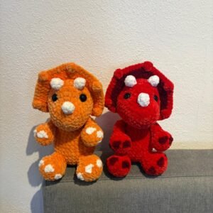Zwei gehäkelte Triceratops-Amigurumi (orange und rot) mit weißen Hörnern und schwarzen Augen, sitzend nebeneinander auf einem grauen Sofa.