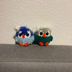 wei gehäkelte Eulen mit großen Glitzeraugen, orangefarbenem Schnabel und Füßen sowie kleinen weißen Federpuscheln, sitzend auf einem grauen Sofa.